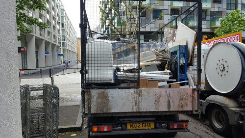 A flatbed truck parked on a city street corner in Dulwich, loaded with a variety of discarded household and office rubbish, including white office chairs, wooden furniture, and cardboard boxes. The truck's cargo area is partially enclosed by a metal cage and shows signs of rust and weathering. Behind the truck, modern apartment buildings with large windows and balconies are visible, along with a black metal fence on the sidewalk. The street surface appears to be paved, and a pedestrian crossing signal is visible at the intersection. The scene suggests a professional rubbish removal service, such as Office Clearance Dulwich, preparing to dispose of waste from an office or property clearance within the local Dulwich area.
