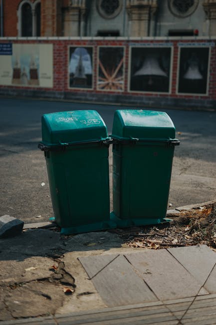 A collection of overflowing waste bins and scattered rubbish on a paved sidewalk in front of a commercial building. The scene includes a large grey mixed paper and card bin with its lid open, revealin