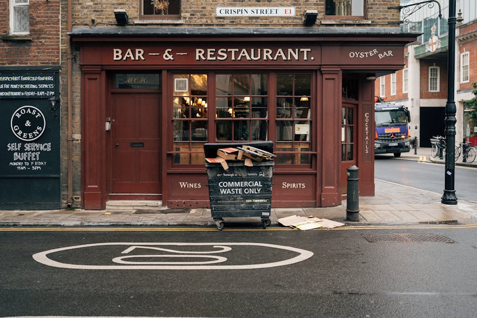 A black commercial waste bin with white lettering reading 'COMMERCIAL WASTE ONLY' is positioned on the sidewalk in front of a restaurant with large glass windows and a maroon exterior. The bin contain