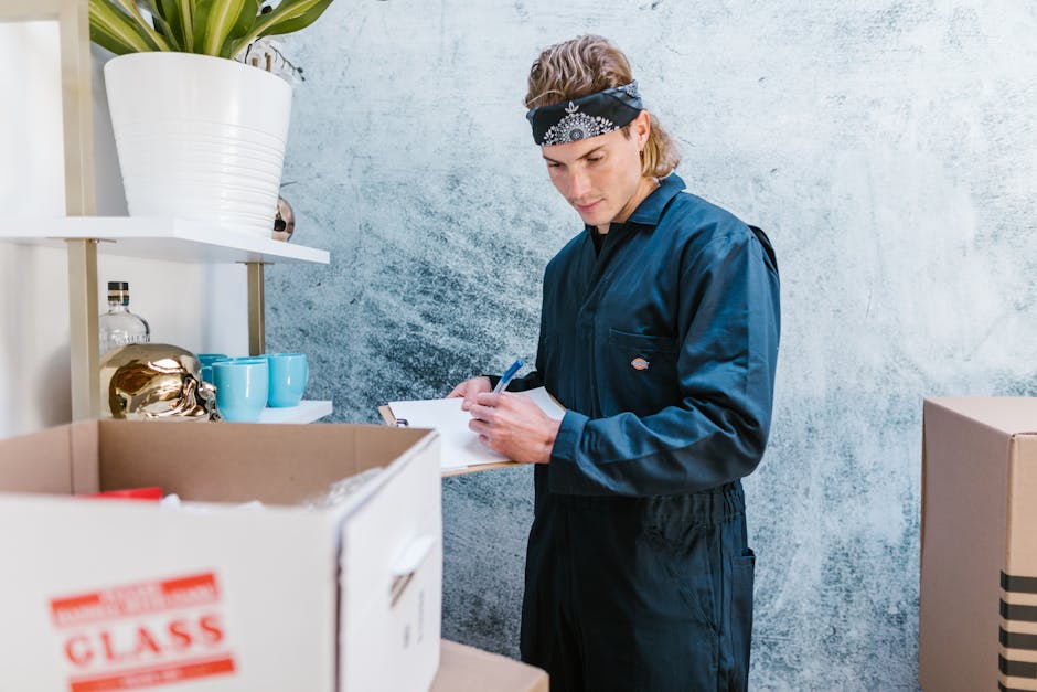 A young man with shoulder-length blonde hair wearing a black bandana and navy-blue jumpsuit is standing indoors against a light, textured wall, examining a notepad with a ballpoint pen. In front of hi